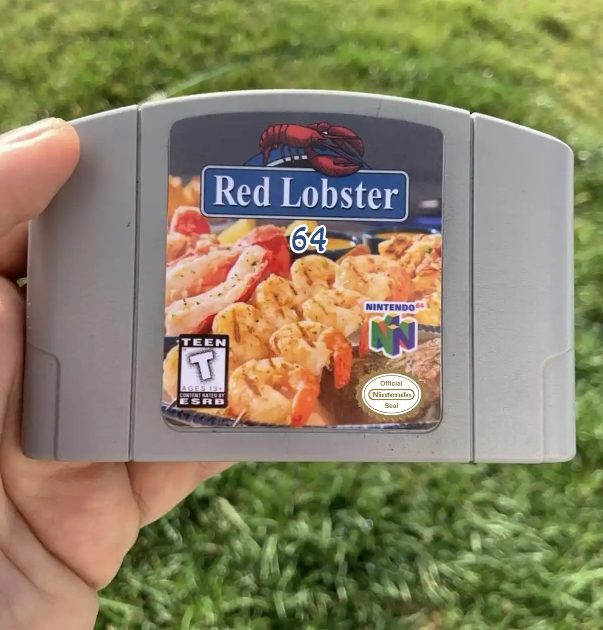 A hand holds a Nintendo 64 game cartridge with a custom label that reads 'Red Lobster 64', featuring an image of grilled…