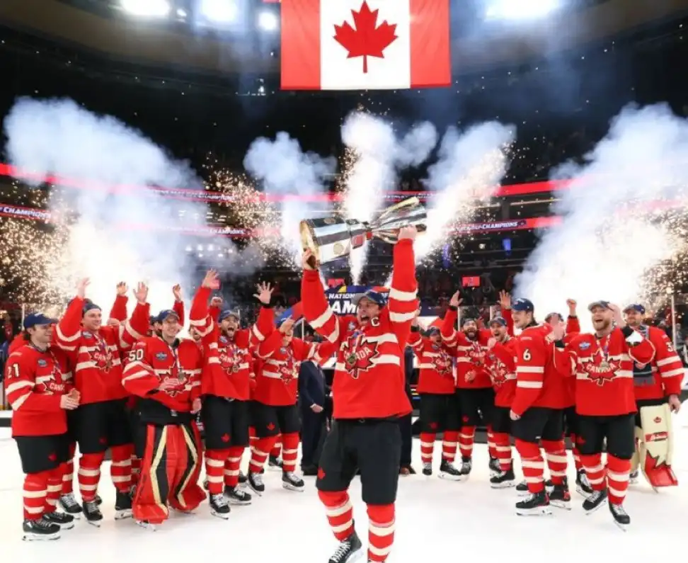 A jubilant Canadian ice hockey team in red jerseys celebrates on the ice, holding a large silver trophy aloft while…