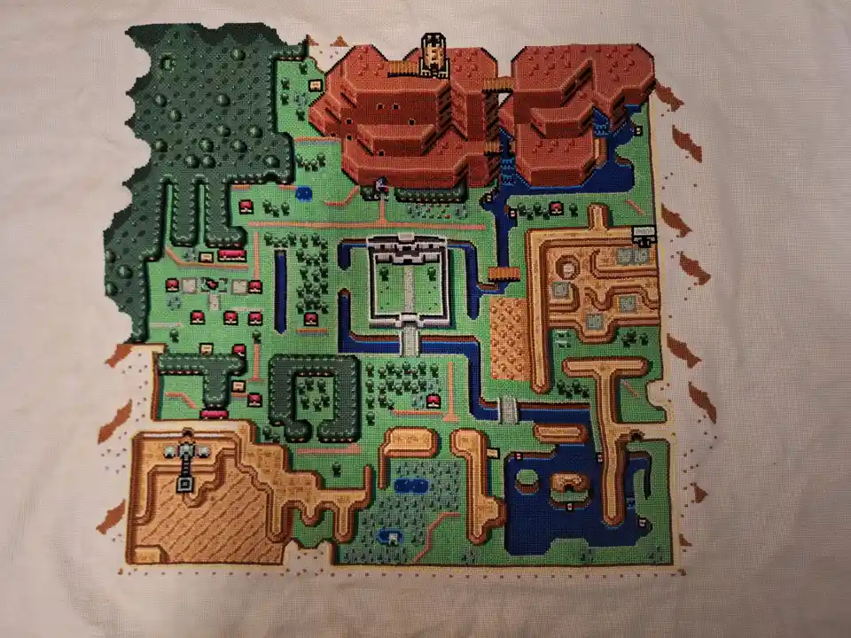 a cross-stitch artwork depicting the pixelated map of the Pokémon Red and Blue game world, stitched onto fabric with green…