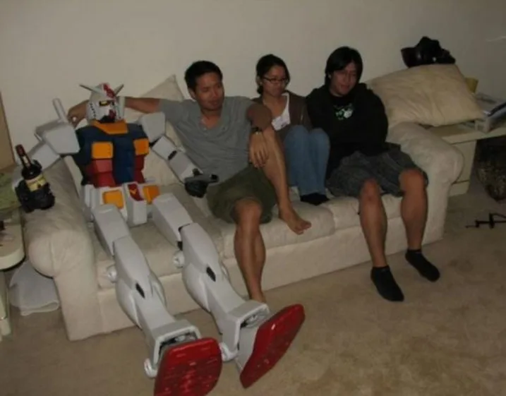 Three people are sitting on a beige couch in a living room, with a large, white and red robot model (resembling a Gundam)…