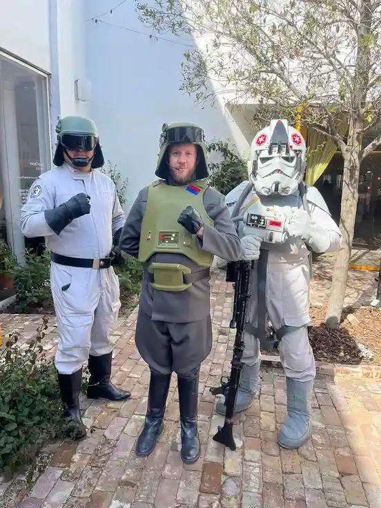 Three men in Star Wars costumes stand side-by-side outdoors on a brick path. The man on the left wears a white uniform with…