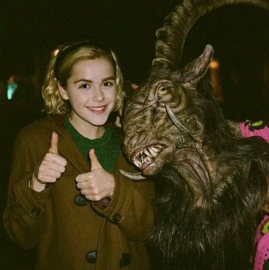 a young woman with blonde hair in a brown coat gives two thumbs up while standing next to a person in a detailed, monstrous…