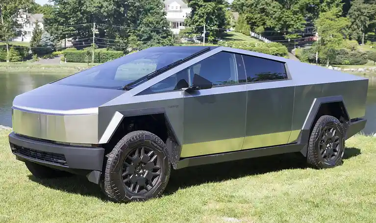 A silver Tesla Cybertruck with a distinctive angular, futuristic design is parked on green grass beside a calm body of…