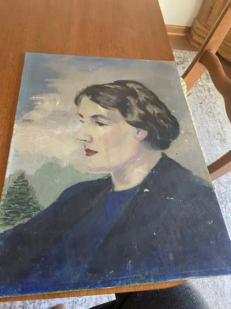 A visibly worn, oil-painted portrait of a woman in profile, wearing a dark blue top, rests on a wooden table. The painting…