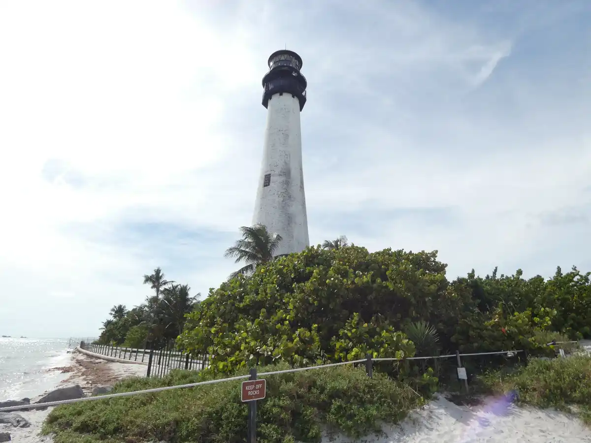 A tall white lighthouse stands on a sandy beach surrounded by lush green vegetation and palm trees under a partly cloudy…
