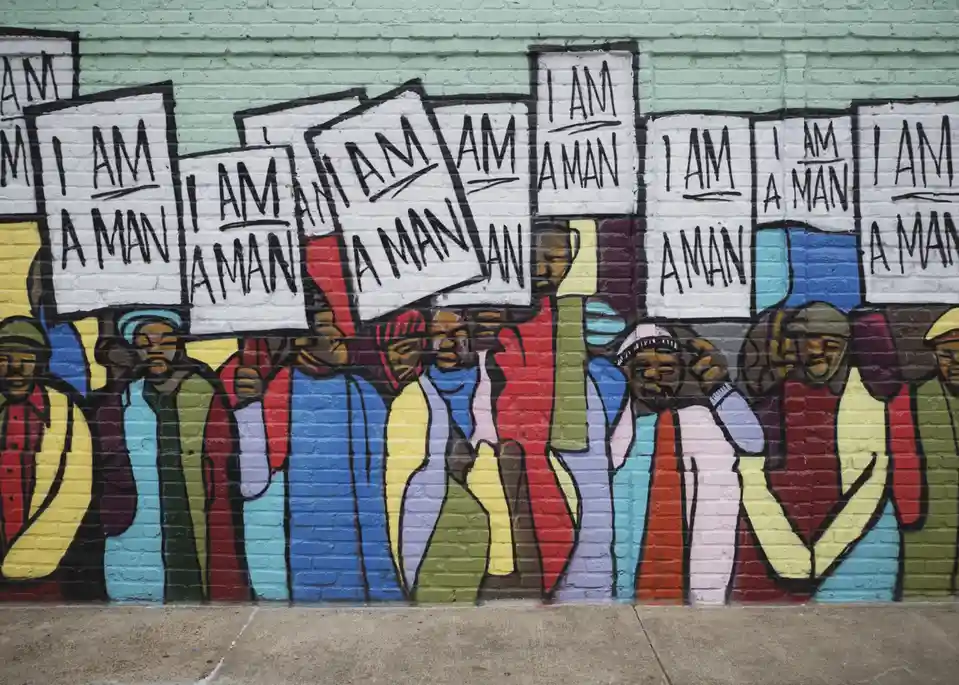 A mural depicts a line of stylized, colorful figures holding up signs that read 'I AM A MAN' in bold black letters. The…