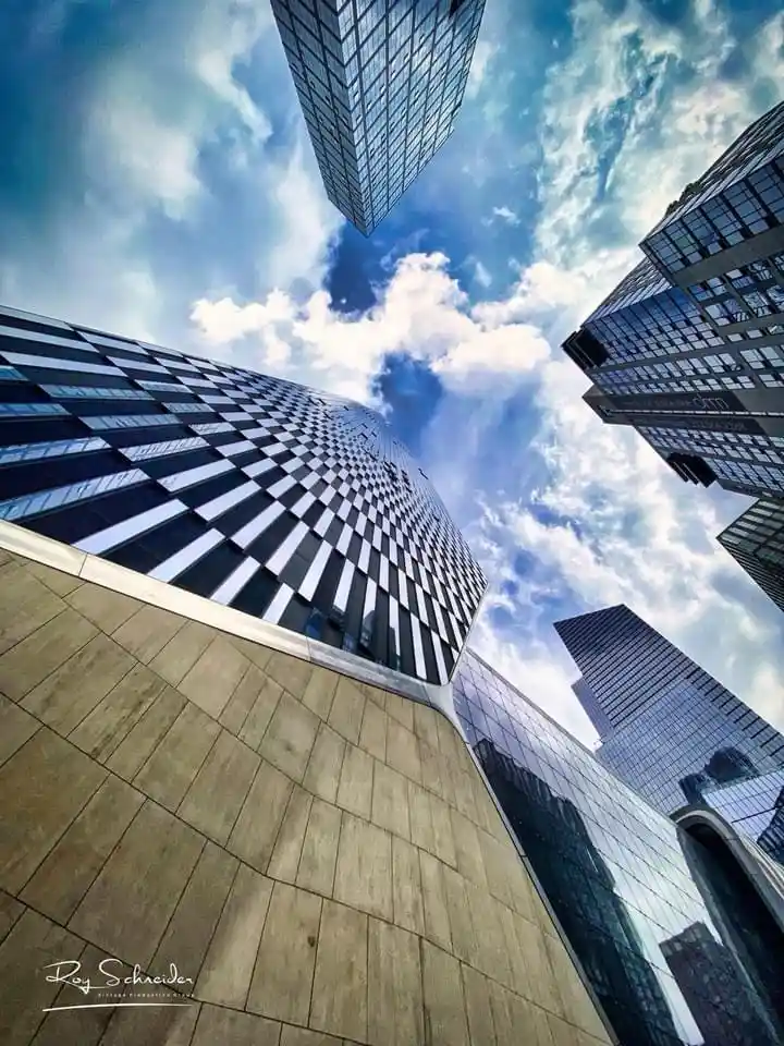 A low-angle photograph shows several towering skyscrapers converging towards a blue sky with white clouds, creating a…