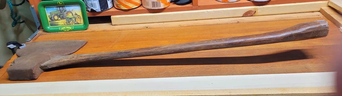 A rusty, old-fashioned axe with a long wooden handle rests on a wooden workbench. In the background, a green John Deere tin…