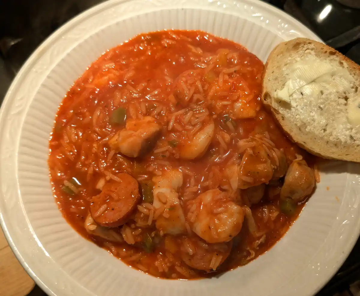 A close-up photo shows a white bowl filled with jambalaya, a dish with rice, shrimp, sausage, and vegetables in a reddish…