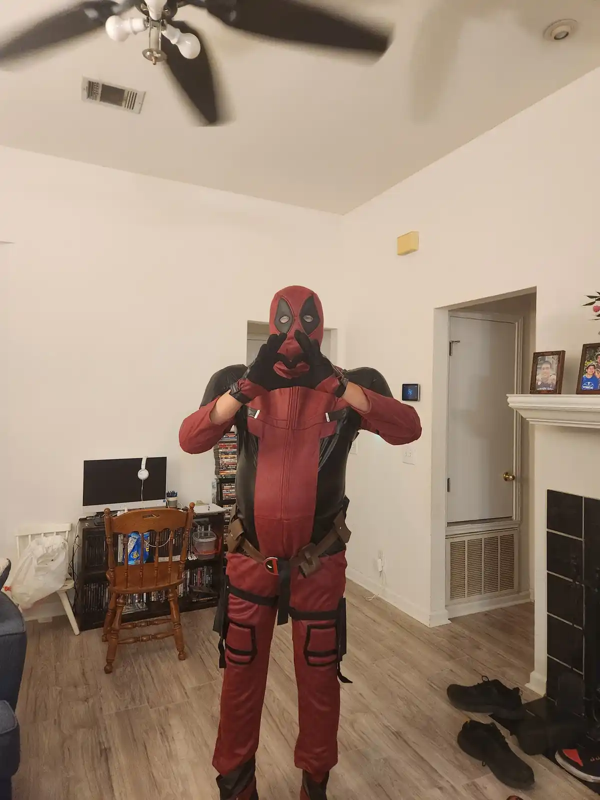 A person in a full Deadpool costume stands in a living room, making a heart shape with their gloved hands near their face…
