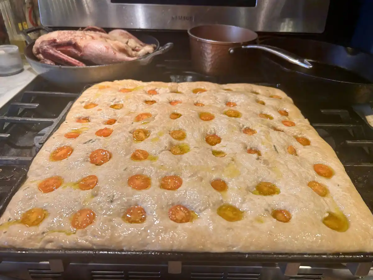 A large, rectangular focaccia bread dough topped with halved cherry tomatoes and olive oil sits on a baking sheet on a…