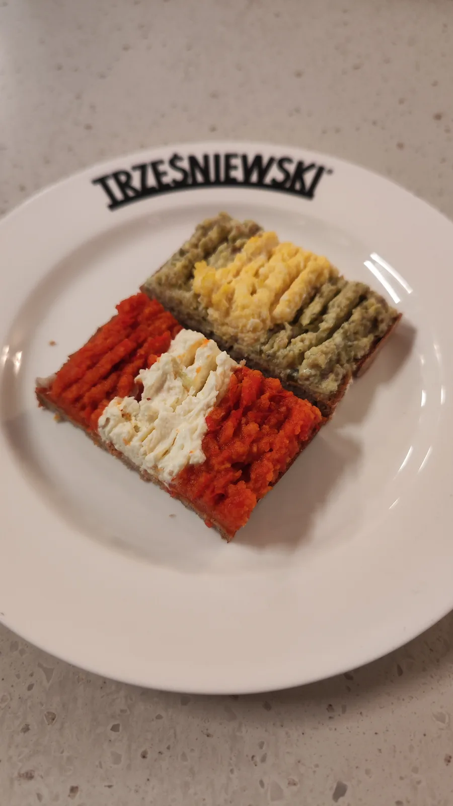 A white plate with the brand name 'TRZESNIEWSKI' printed on its rim holds two square pieces of bread topped with colorful…