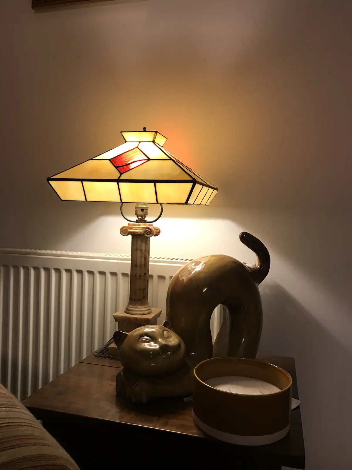 A warmly lit stained-glass lamp sits on a wooden table next to a ceramic cat statue and a candle holder. The statue is…