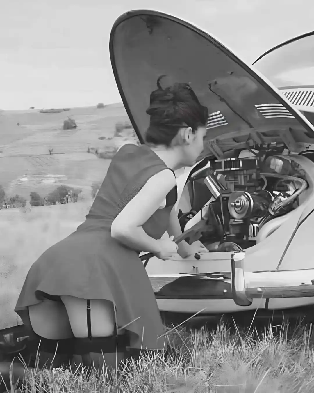 A black-and-white photo shows a woman in a 1950s-style dress and stockings kneeling beside a vintage car with its hood open…