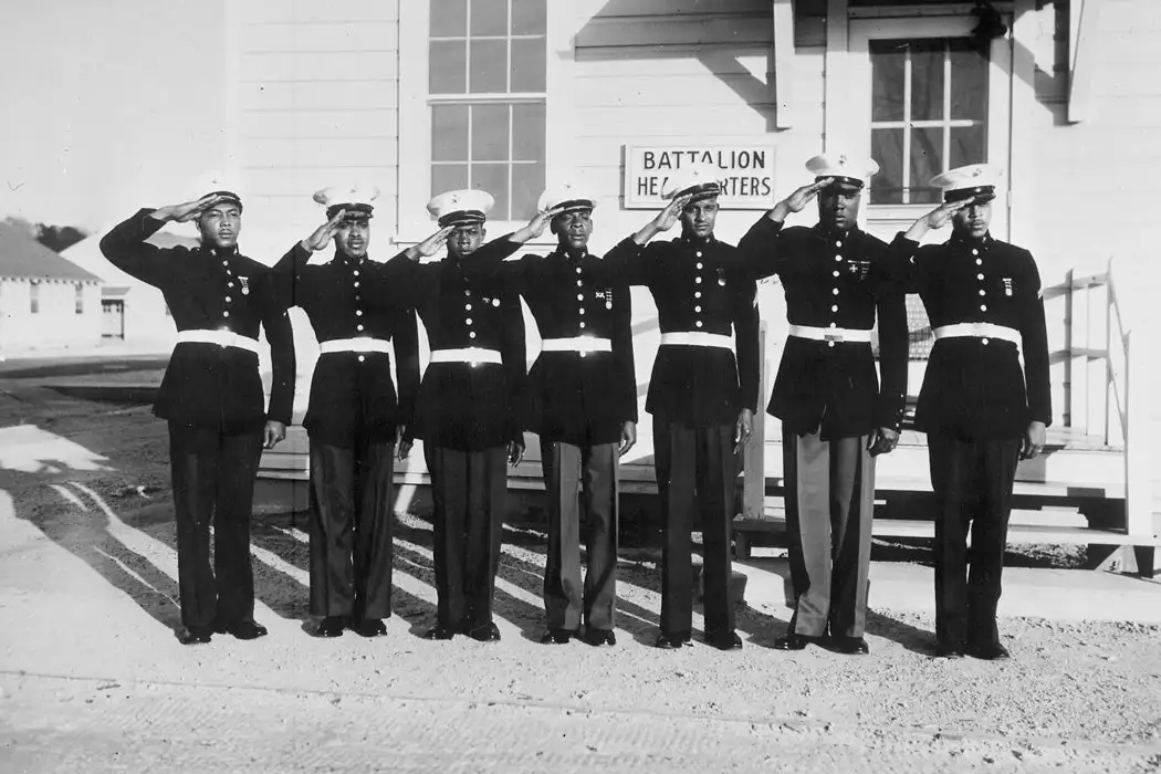 A black-and-white historical photo shows seven Black U.S. Marines in dress uniforms saluting in front of a building labeled…
