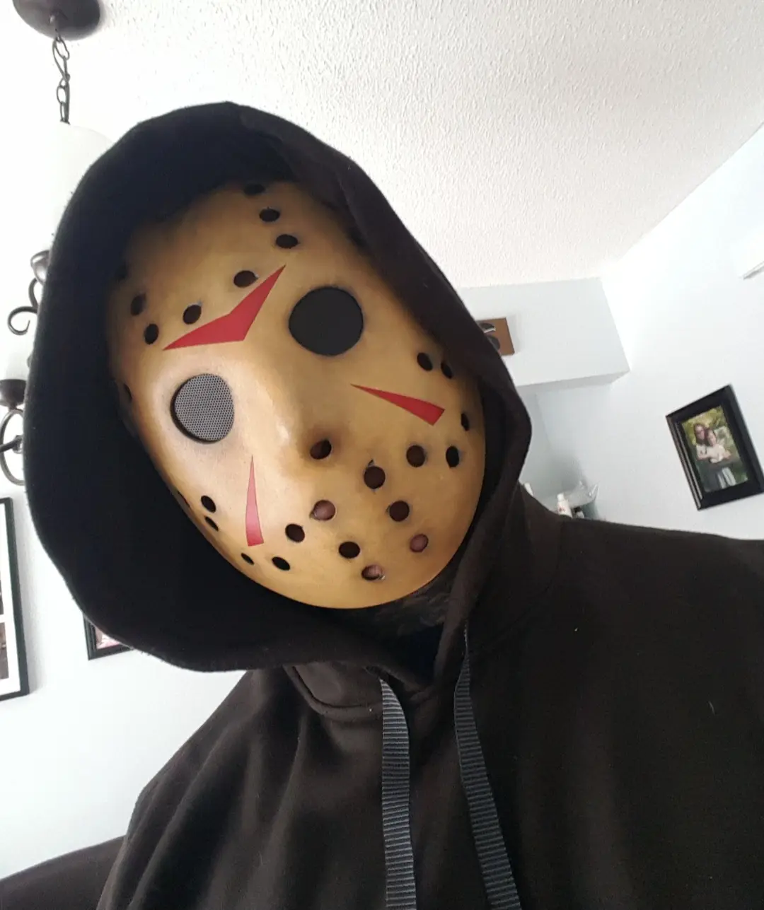 A person wearing a brown hoodie with the hood up, obscuring their hair and neck, and a tan hockey mask with black eye holes…
