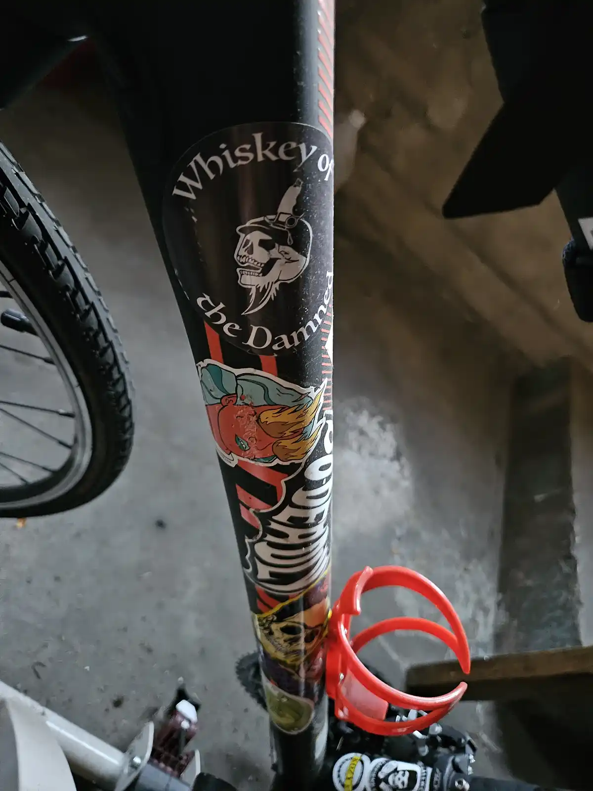 A close-up of a black bicycle frame covered in stickers, including a prominent 'Whiskey of the Damned' logo with a skull…