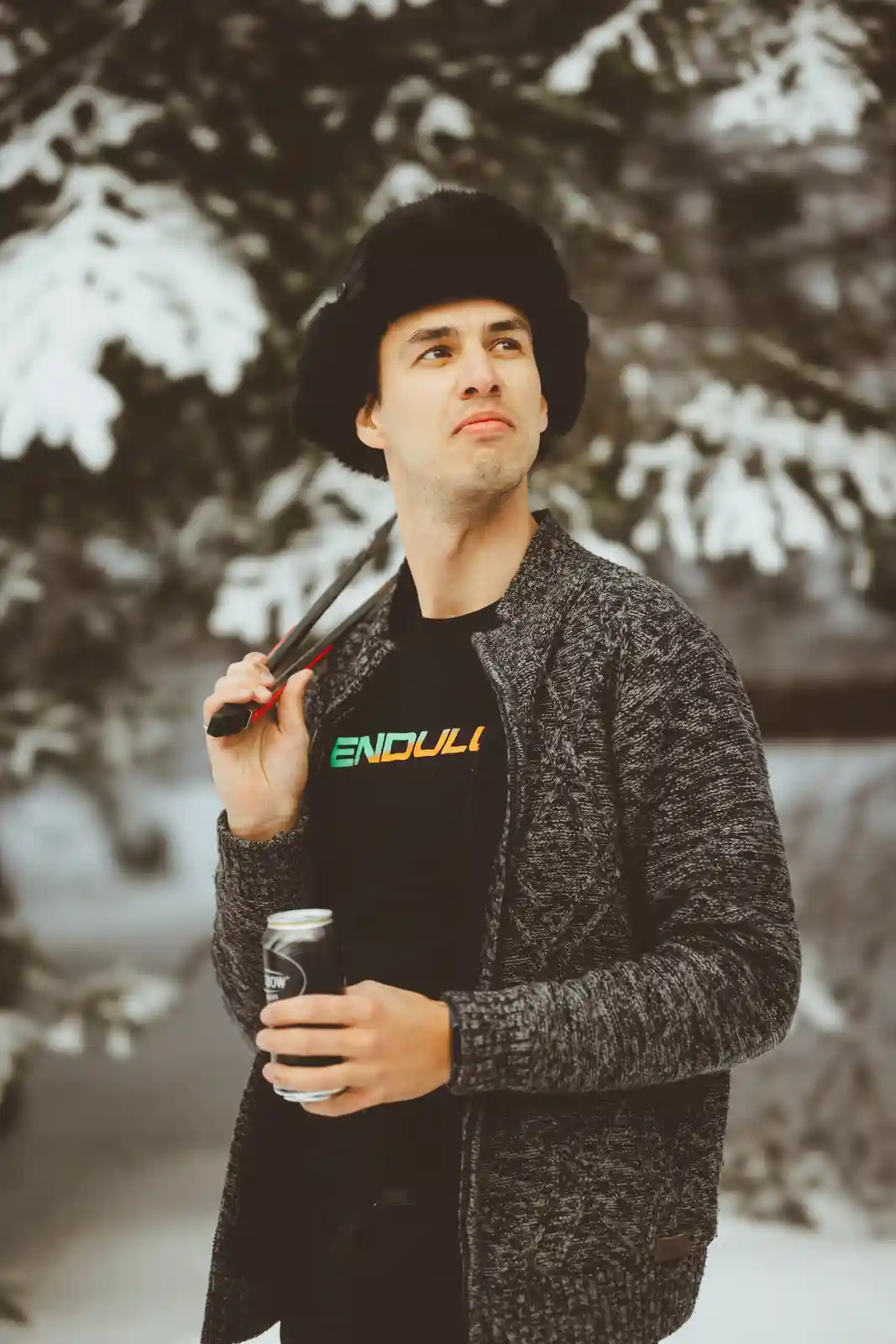 A man in a snowy forest wearing a black fur hat and grey knit cardigan, holding a beer can and a pair of ski poles, looks…