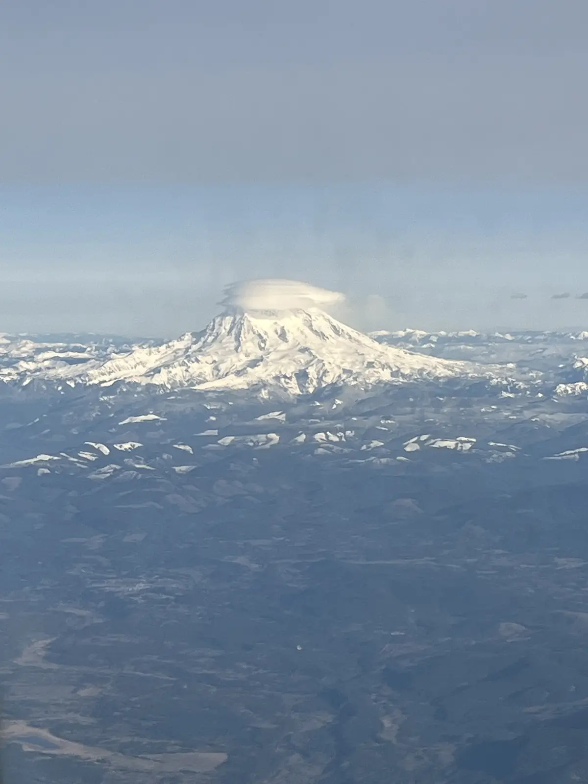 An aerial view of a snow-capped mountain, likely Mount Rainier, with a distinct, flat-topped cloud formation hovering…