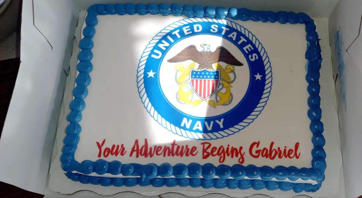 A rectangular white cake with blue piped borders features the United States Navy seal in the center, with red icing text…