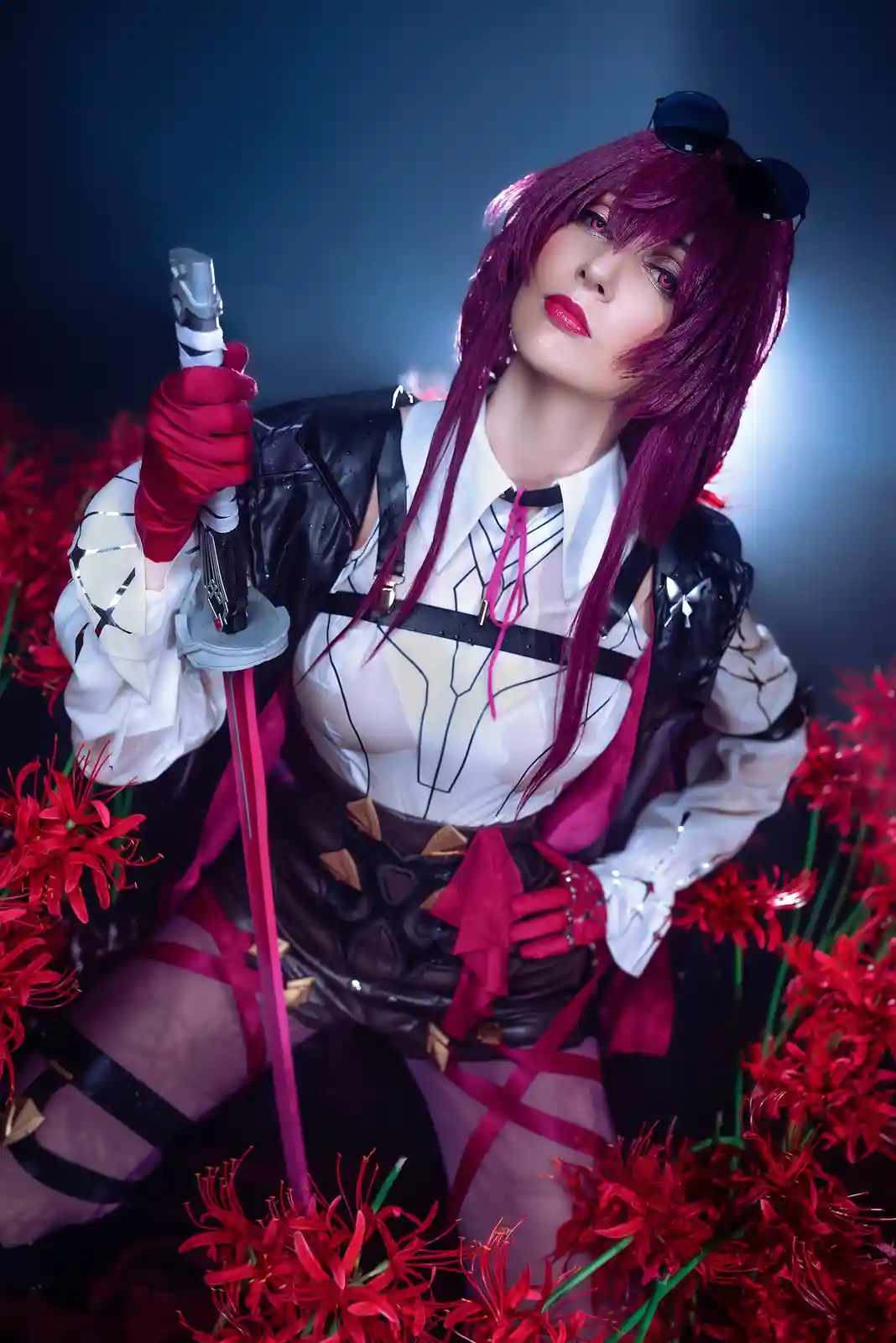 A person in cosplay with long purple hair, red gloves, and a white and black outfit holds a sword while sitting among red…