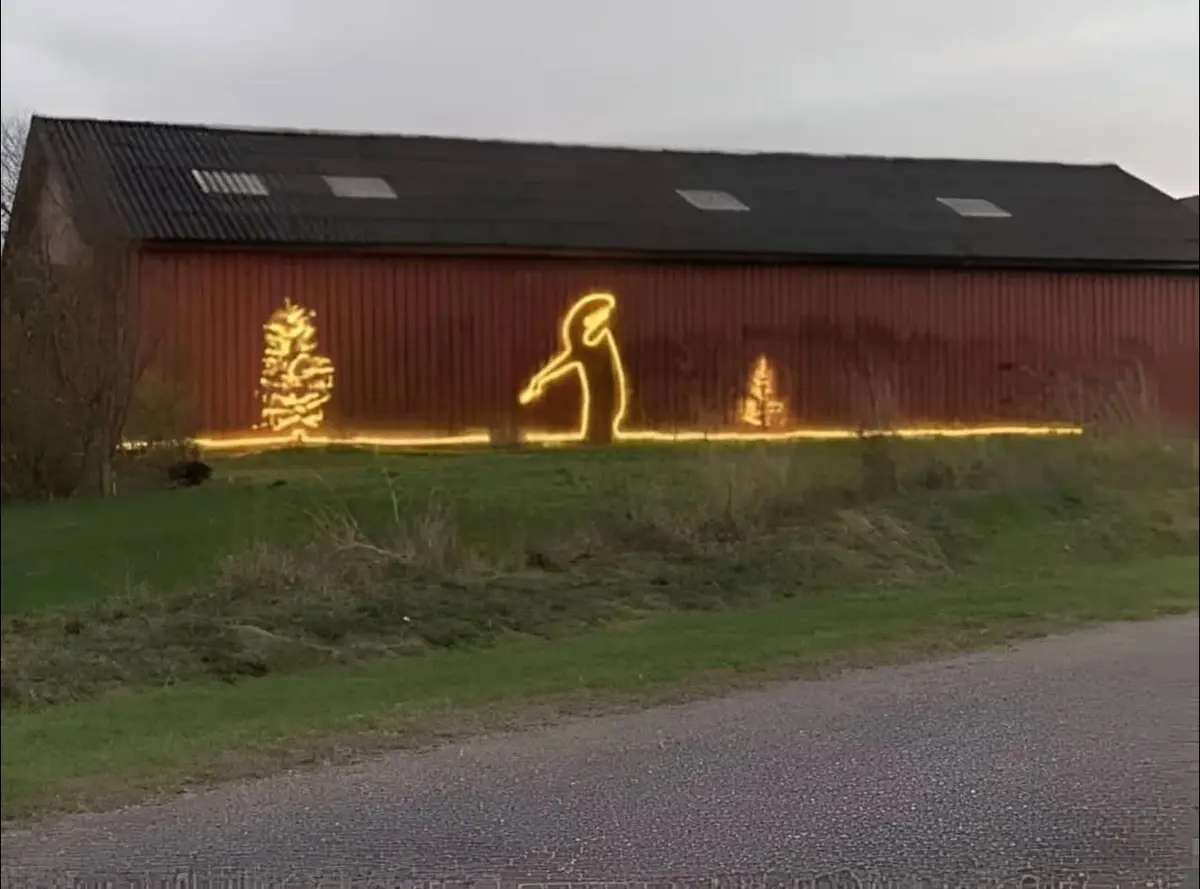A red barn with festive yellow string lights outlining two Christmas trees and a large, stylized figure of a person in a…