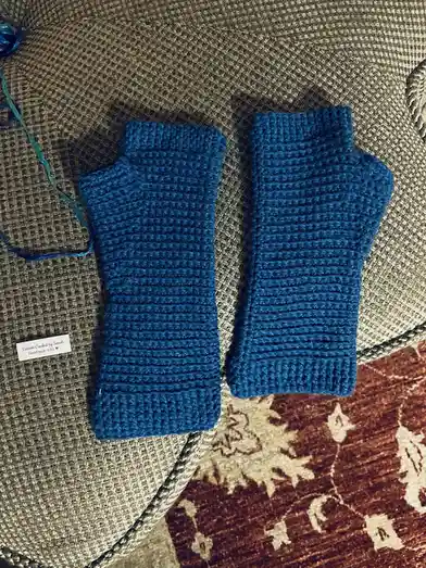 Two blue crocheted fingerless gloves lie side by side on a textured beige surface, with a small white tag reading 'Custom…