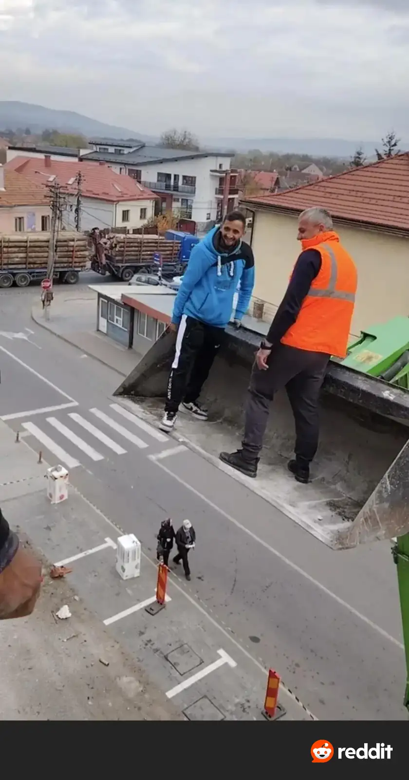 Two men stand in the bucket of a construction crane overlooking a street with pedestrians below. One man in a blue hoodie…