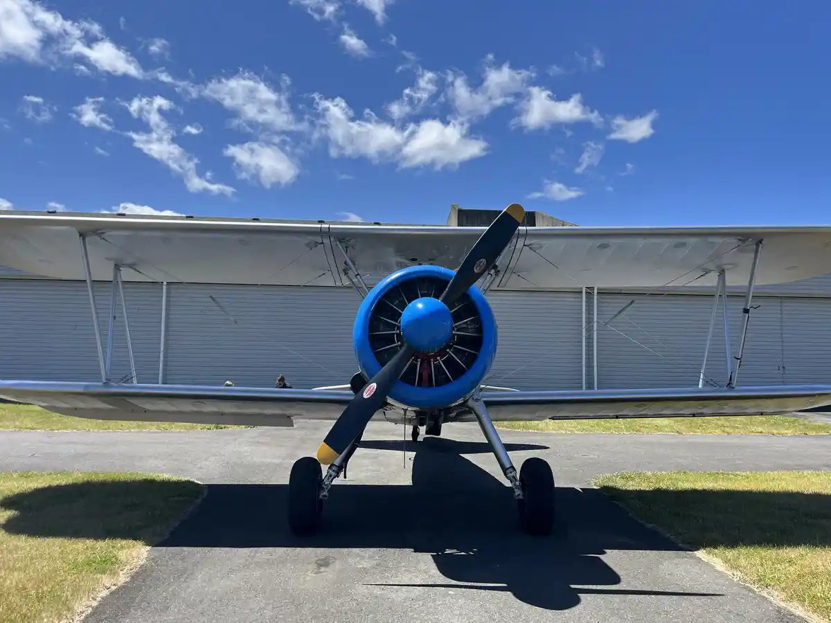 A front-facing photo of a silver biplane with a bright blue engine cowling and black propeller blades tipped with yellow…