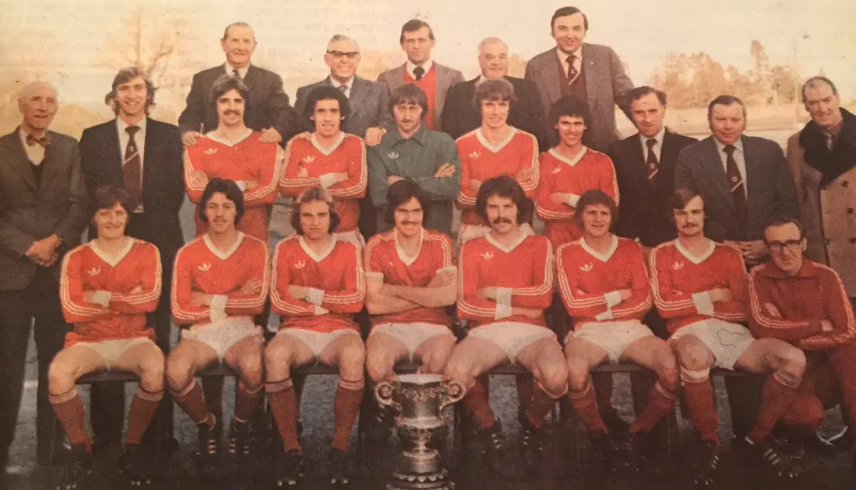 A vintage team photo shows a group of men in red and white soccer uniforms posing with a large silver trophy placed between…