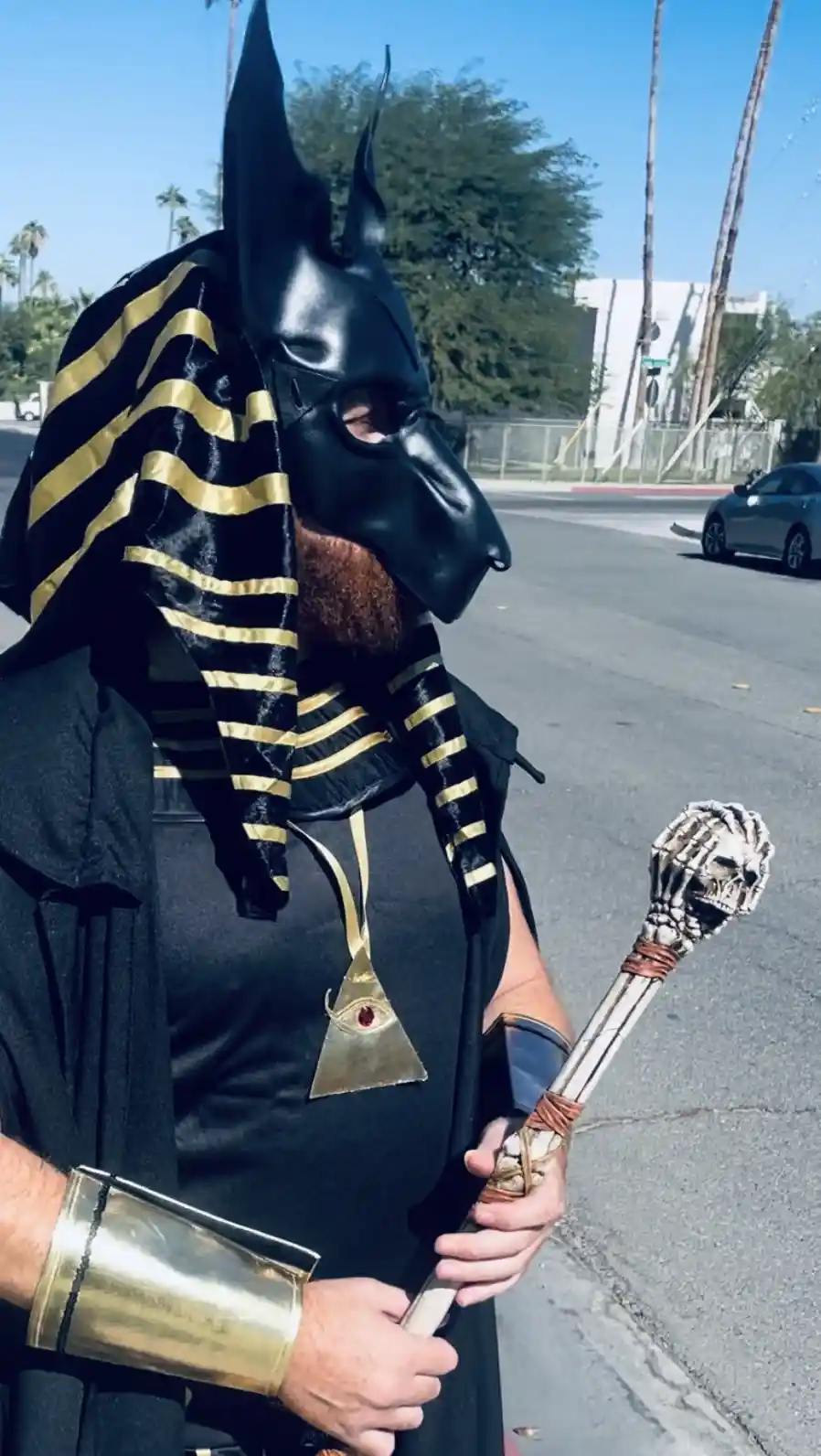 A person wearing a black Anubis dog-headed mask with a black-and-gold striped headdress stands on a street, holding a staff…