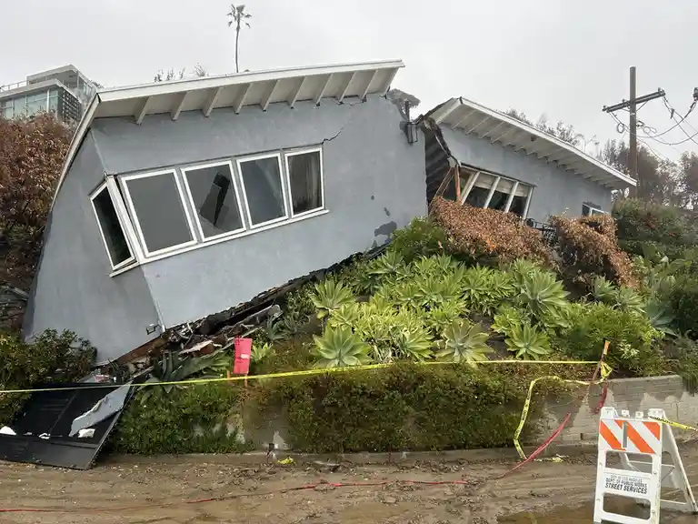 A light blue house is visibly tilted and damaged, sliding down a hillside with broken windows and exposed framing…
