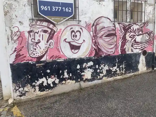 A weathered wall features a faded graffiti mural of four cartoonish faces with exaggerated expressions, painted in pink and…