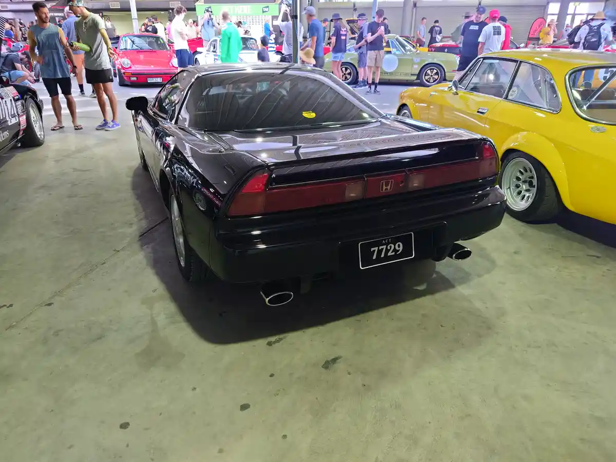 A black Honda NSX is parked at a car show, viewed from the rear, with its license plate reading 'ACT 7729'. Surrounding it…