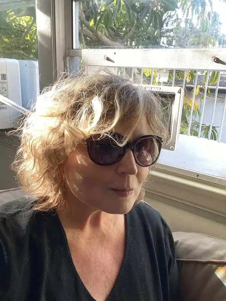 A woman with curly blonde hair and sunglasses sits indoors near a window, with a 'CAT MATE' pet door mounted on the window…