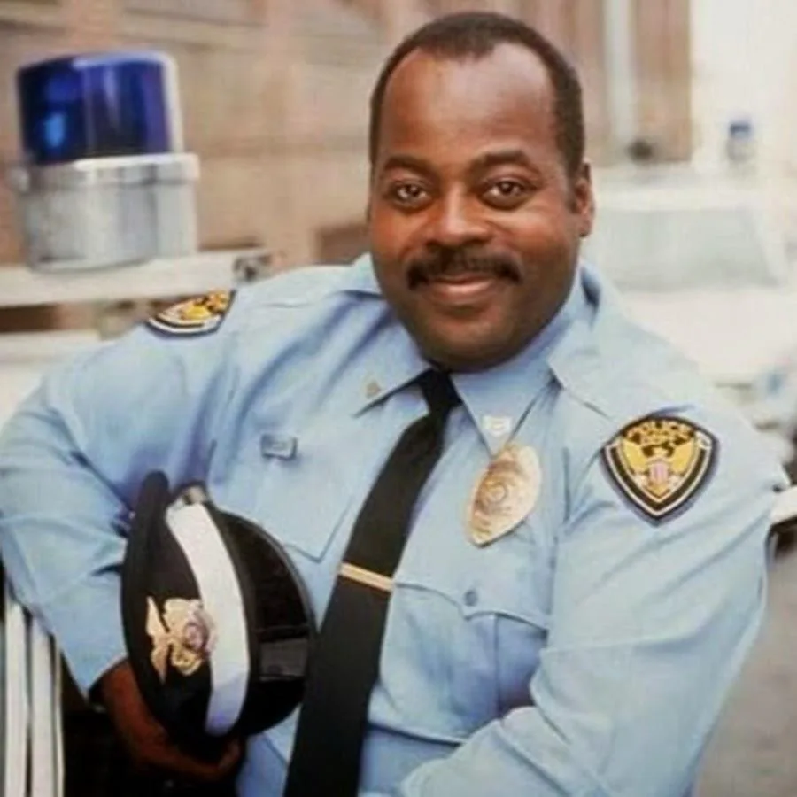 A smiling Black man in a light blue police uniform with a badge and shoulder patch, holding a police hat. A blue police…