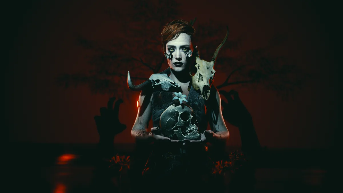 A person with short red hair and butterfly makeup holds a human skull, flanked by two animal skulls, against a dark red…