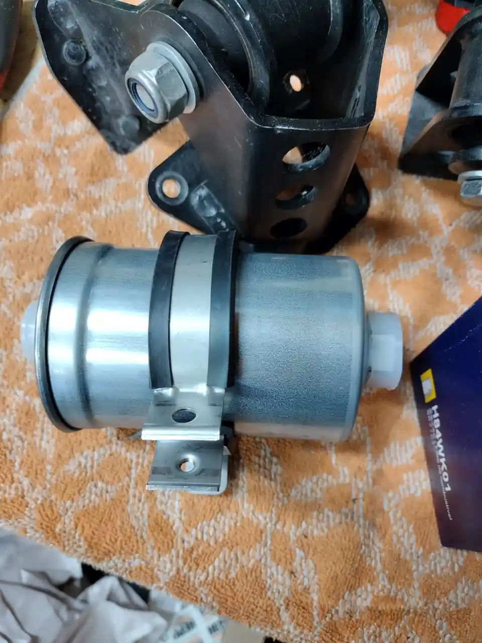 A close-up photo shows a new-looking silver cylindrical fuel filter with black rubber gaskets and mounting brackets, resting…