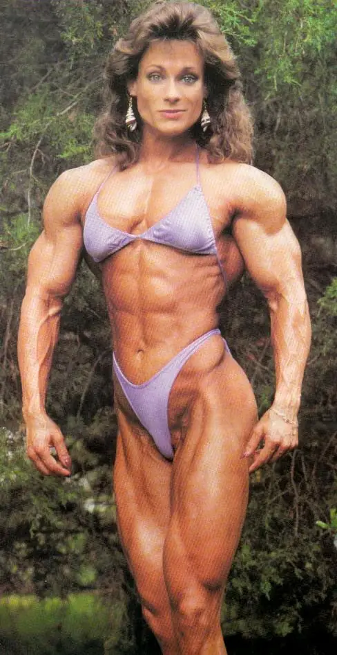 A woman with extremely muscular, defined physique poses outdoors in a purple bikini, showcasing her prominent abdominal…