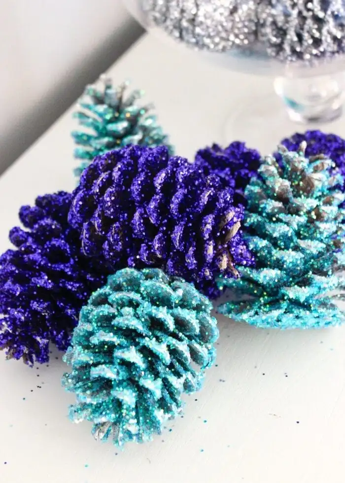 A close-up photo shows several pinecones covered in glitter, with some in vibrant purple and others in teal, arranged on a…