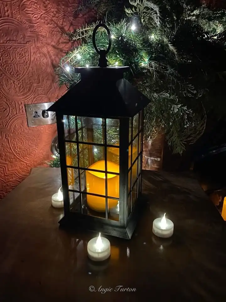 A cozy indoor scene features a black metal lantern with a glowing yellow candle inside, surrounded by three lit white tea…