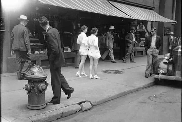 A black-and-white photo from the 1940s shows a man in a suit walking past a fire hydrant on a city sidewalk. In the…