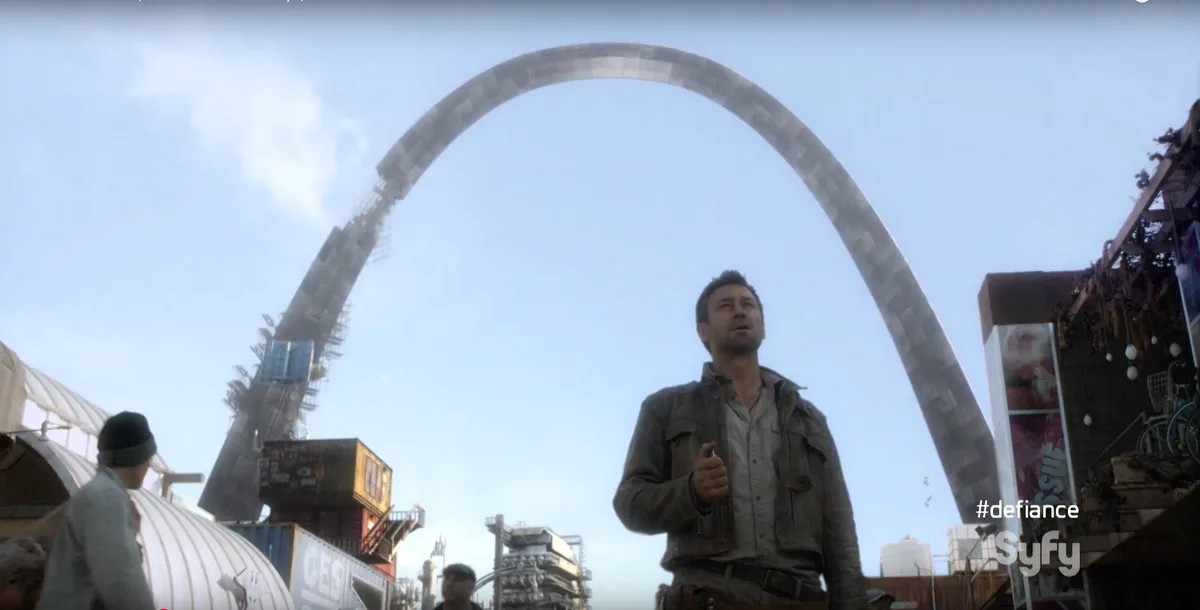 A man in a worn jacket stands looking up in awe at a massive, futuristic, curved metal arch dominating a gritty…