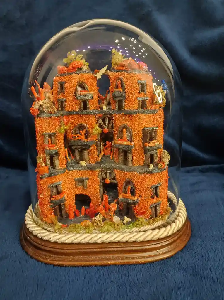 A miniature, multi-story building model, entirely covered in orange coral and sea creatures, is displayed under a glass dome…