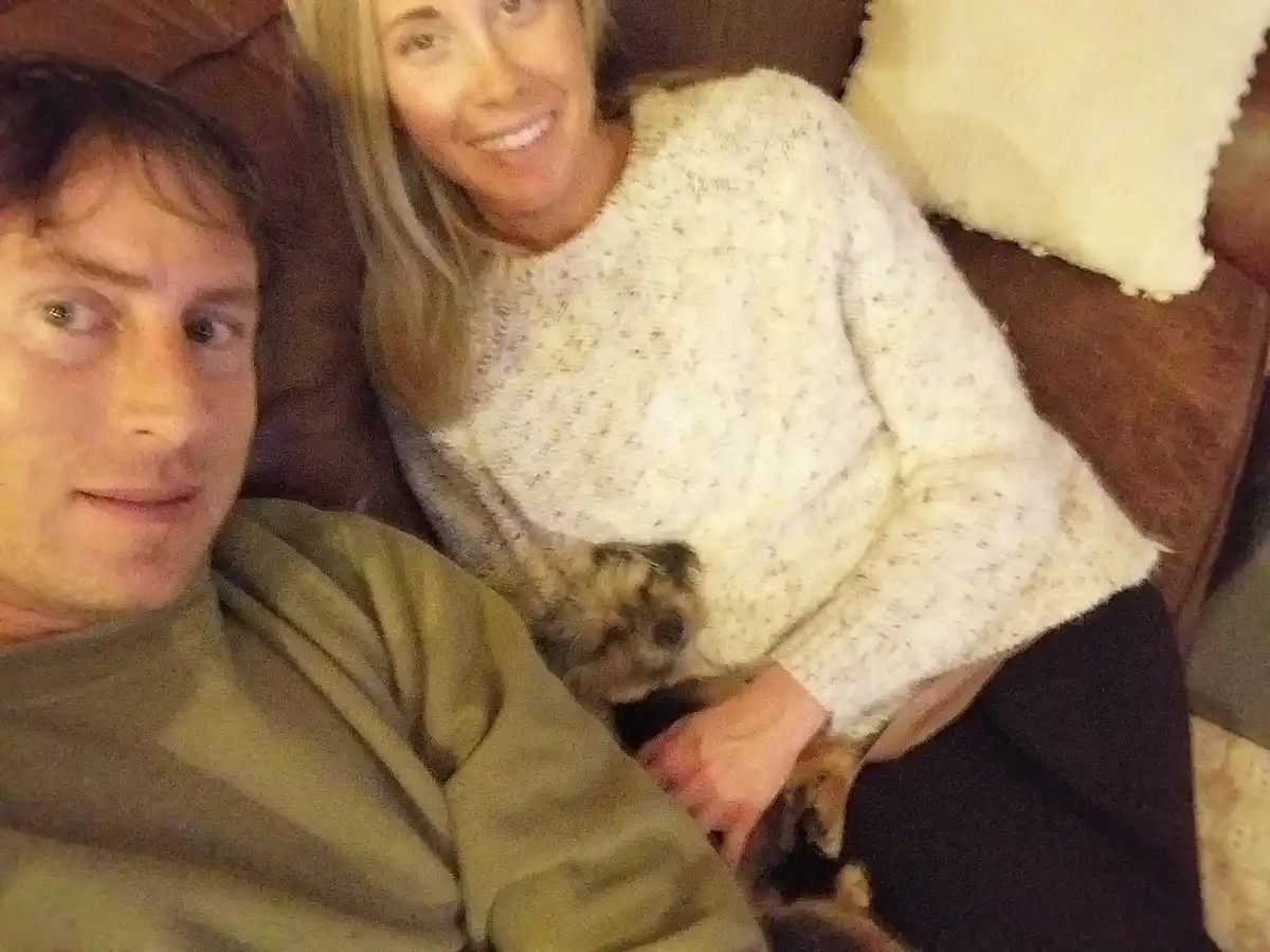 A selfie-style photo shows a man and a woman sitting on a brown couch, smiling at the camera. The woman, wearing a speckled…