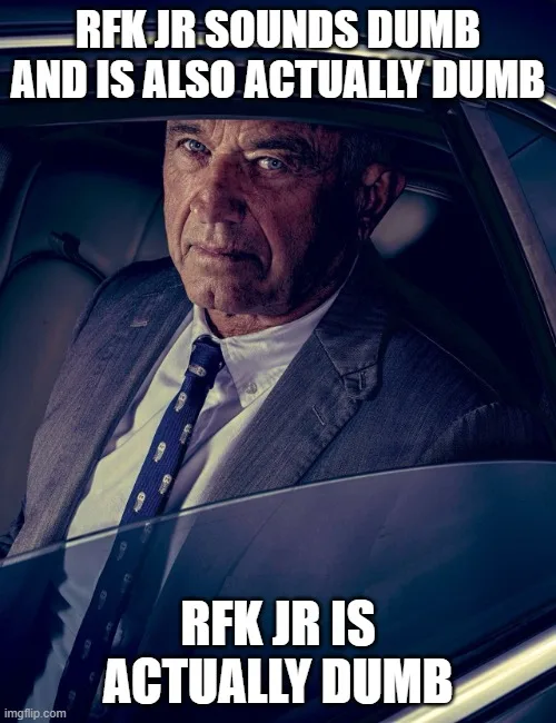 A close-up photo of a man in a suit and tie sitting inside a car, looking intensely at the camera. The image has two lines…