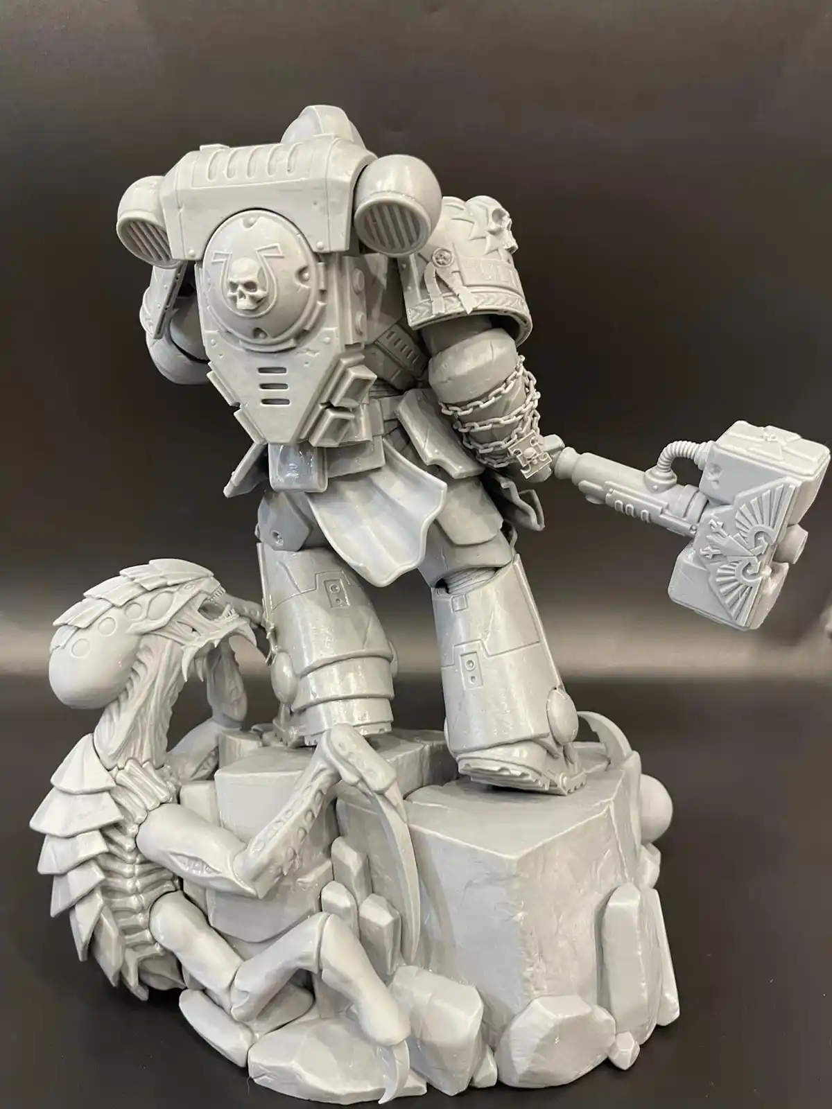 A grey, unpainted miniature of a heavily armored space marine stands triumphantly over a defeated alien creature, both…