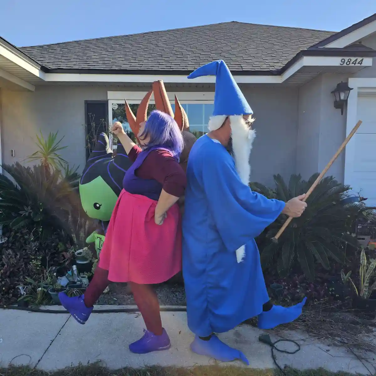 Two people stand back-to-back on a residential sidewalk in front of a house, dressed as characters from 'The Legend of…