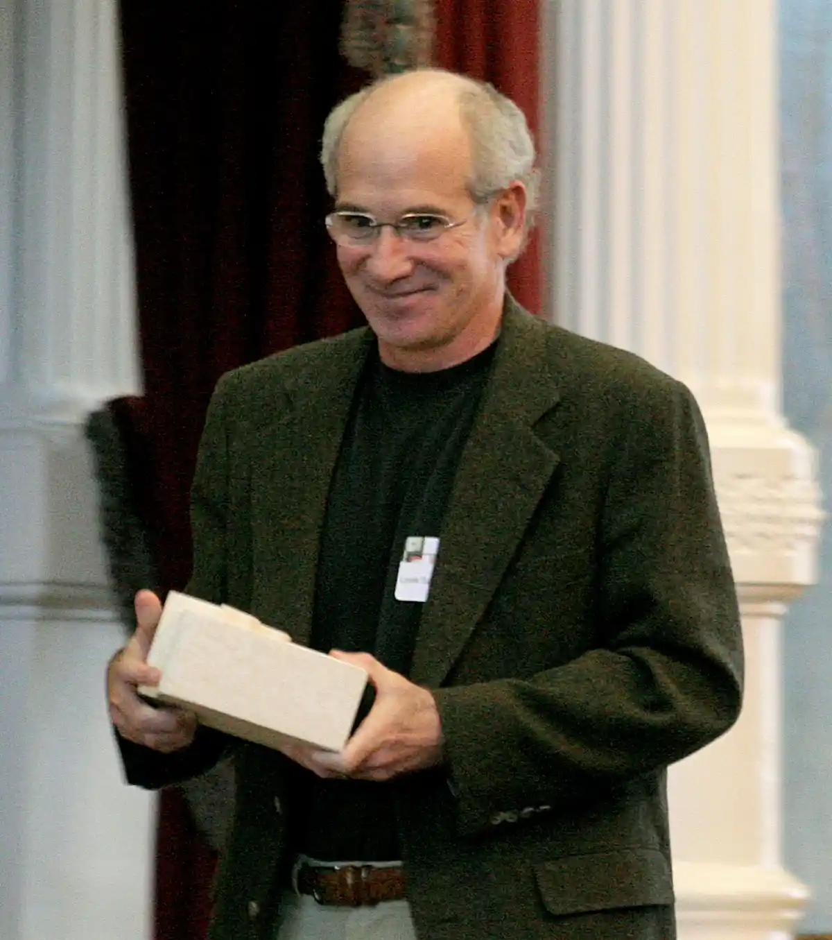 a man with glasses and a name tag smiles while holding a rectangular box, standing in front of columns and red curtains. the…