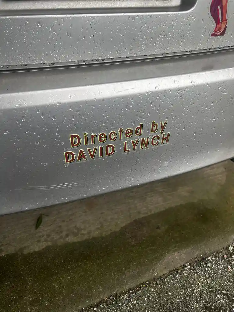 A close-up of a silver car's side panel covered in rain droplets, featuring a sticker that reads 'Directed by DAVID LYNCH'…